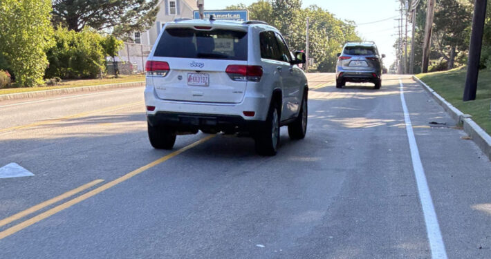 Cyclist's view of a two-way road with a two-way turn lane and narrow should with a crrb, cyclist riding in the right tire track, motorist straddle passing.